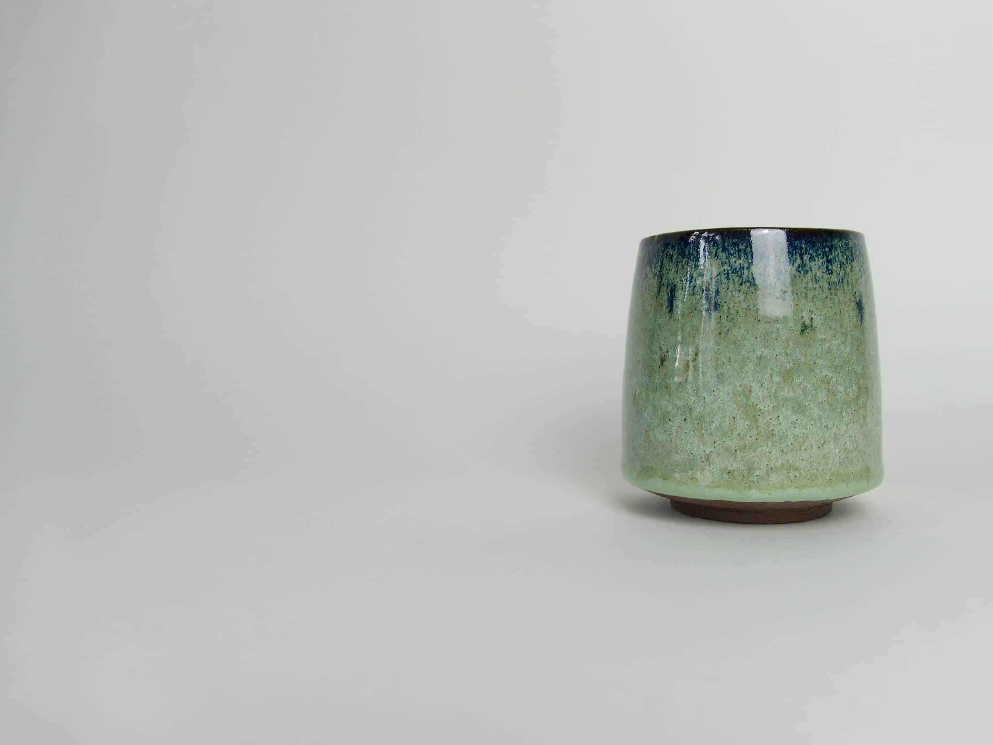 Handmade stoneware cup with a pale green glaze and dark blue rim on a light background.