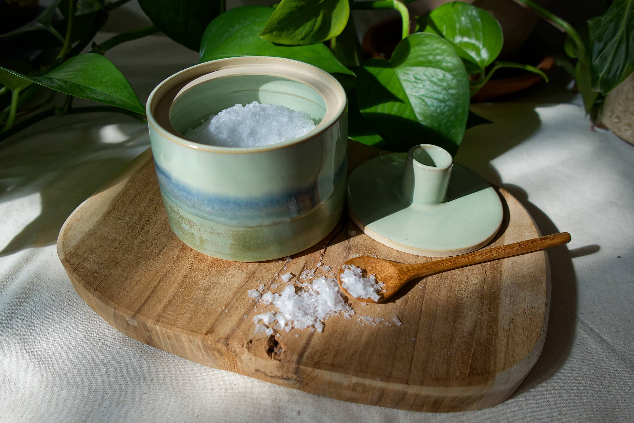 Pale green glazed ceramic salt jar with its lid and a wooden spoon on a serving board.